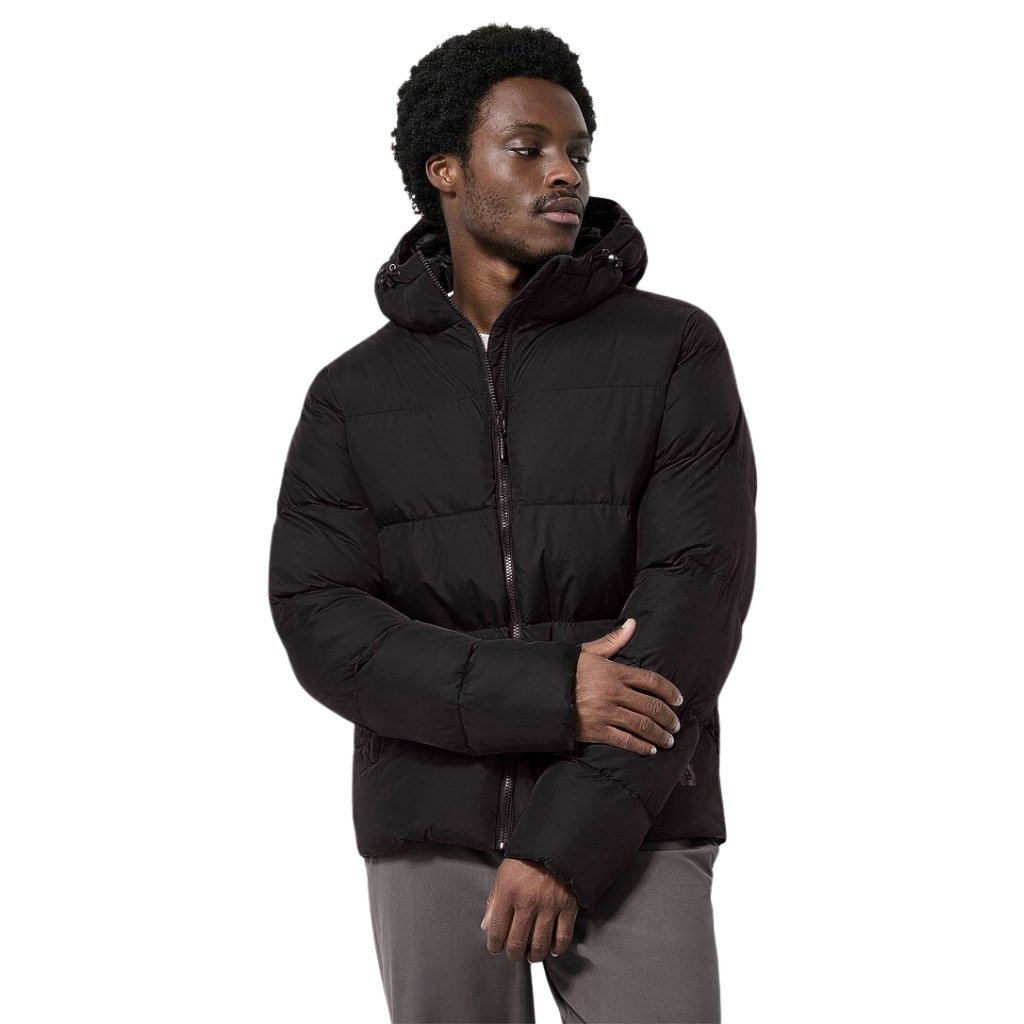 : A man with dark curls wearing a black puffer jacket with a hood, standing against a white background, arms crossed and looking to the side.