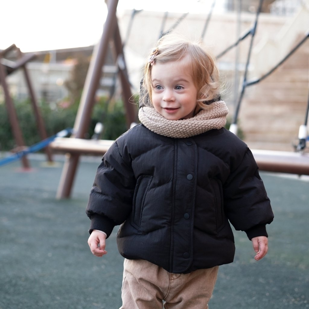 A young child wearing a black down  jacket, beige pants, and a tan scarf stands on a green playground surface. Play structures and greenery appear in the background as the child looks slightly to the right with a mild smile.