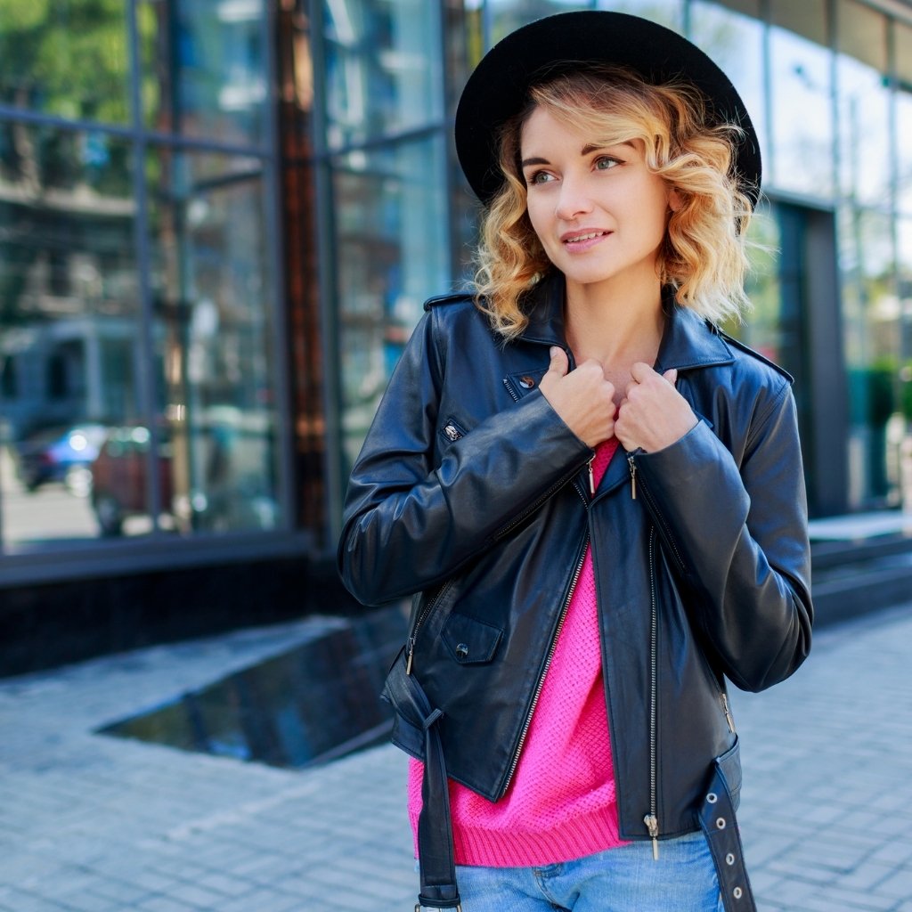 A young woman stands outside a modern glass-front building, wearing a black hat, a black leather jacket, a bright pink sweater, and light blue jeans. She holds the lapels of her jacket with both hands and looks to her right, with the glass facade reflecting the surroundings.