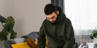 Cropped Hoodie Styles: Trendy, Comfortable & Modern Wear Up A man wearing glasses and a green hoodie leans over a dark bag indoors, reaching inside. He stands near a gray couch with a yellow pillow; a green plant sits behind the couch, and a white desk with a red object is visible on the right.