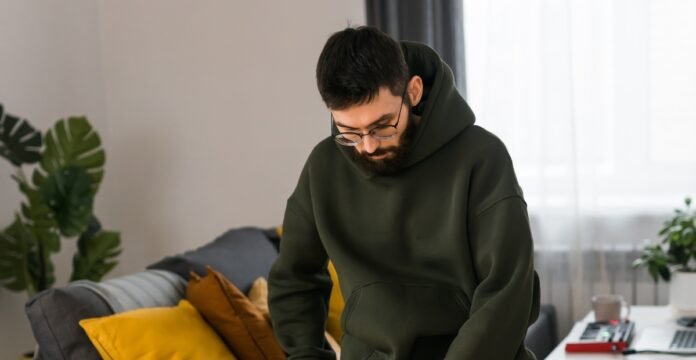 A man wearing glasses and a green hoodie leans over a dark bag indoors, reaching inside. He stands near a gray couch with a yellow pillow; a green plant sits behind the couch, and a white desk with a red object is visible on the right.