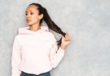 Cropped Plain Hoodie: Minimal Style, Maximum Comfort A portrait of a young woman wearing a light pink hoodie, standing against a gray textured wall. She has dark hair tied in a ponytail and is holding the end of her ponytail with one hand, looking to the side.