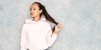 Cropped Plain Hoodie: Minimal Style, Maximum Comfort A portrait of a young woman wearing a light pink hoodie, standing against a gray textured wall. She has dark hair tied in a ponytail and is holding the end of her ponytail with one hand, looking to the side.