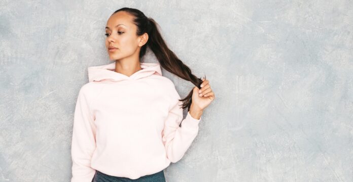A portrait of a young woman wearing a light pink hoodie, standing against a gray textured wall. She has dark hair tied in a ponytail and is holding the end of her ponytail with one hand, looking to the side.