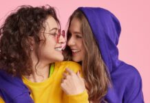 Cropped Zip Hoodie: Trendy Streetwear Style Guide Two young women smiling and embracing against a pink backdrop. The woman on the left has curly brown hair, rose-tinted glasses, a yellow shirt with lime green trim, and a dark purple hoodie. The woman on the right has long brown hair and wears a dark purple hoodie.