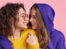 Cropped Zip Hoodie: Trendy Streetwear Style Guide Two young women smiling and embracing against a pink backdrop. The woman on the left has curly brown hair, rose-tinted glasses, a yellow shirt with lime green trim, and a dark purple hoodie. The woman on the right has long brown hair and wears a dark purple hoodie.