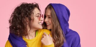Cropped Zip Hoodie: Trendy Streetwear Style Guide Two young women smiling and embracing against a pink backdrop. The woman on the left has curly brown hair, rose-tinted glasses, a yellow shirt with lime green trim, and a dark purple hoodie. The woman on the right has long brown hair and wears a dark purple hoodie.