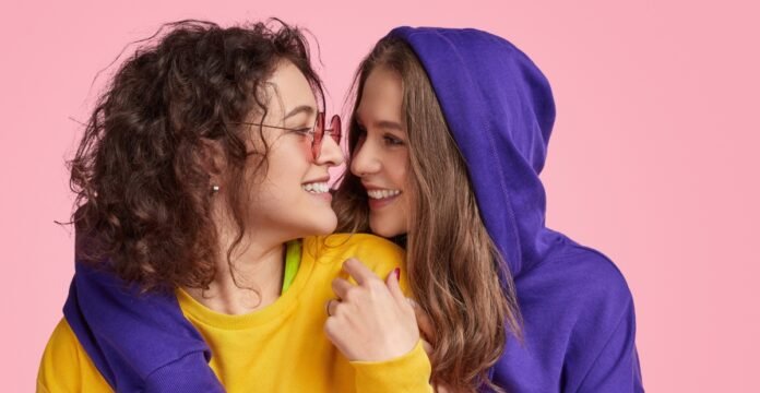 Two young women smiling and embracing against a pink backdrop. The woman on the left has curly brown hair, rose-tinted glasses, a yellow shirt with lime green trim, and a dark purple hoodie. The woman on the right has long brown hair and wears a dark purple hoodie.