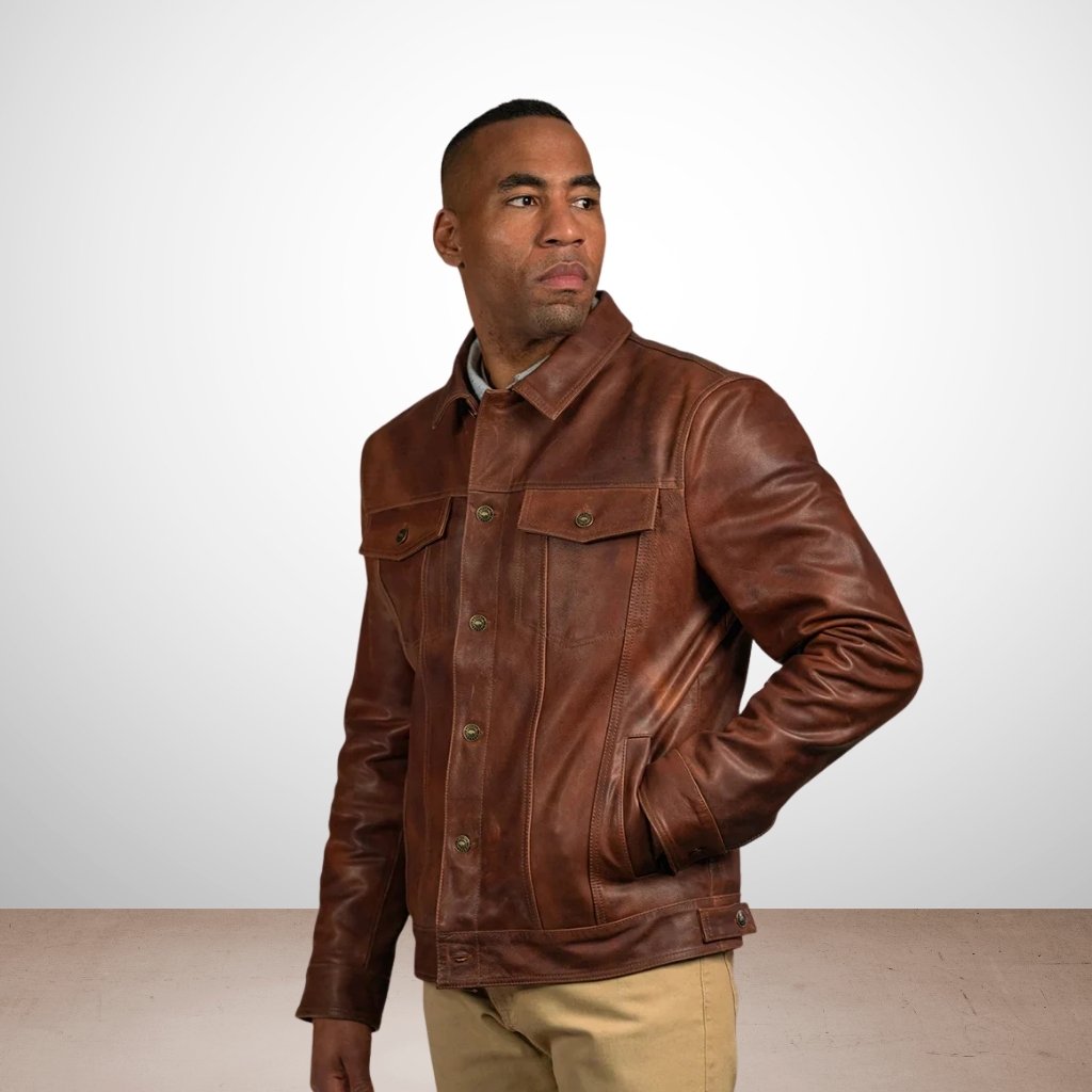 Alt text (for screen readers): An African American man stands against a white backdrop with a tan floor, wearing a brown leather trucker jacket with two chest pockets, button closure, and a collar. He has his left hand in his pocket and is looking to the side.denim Style Leather Jacket