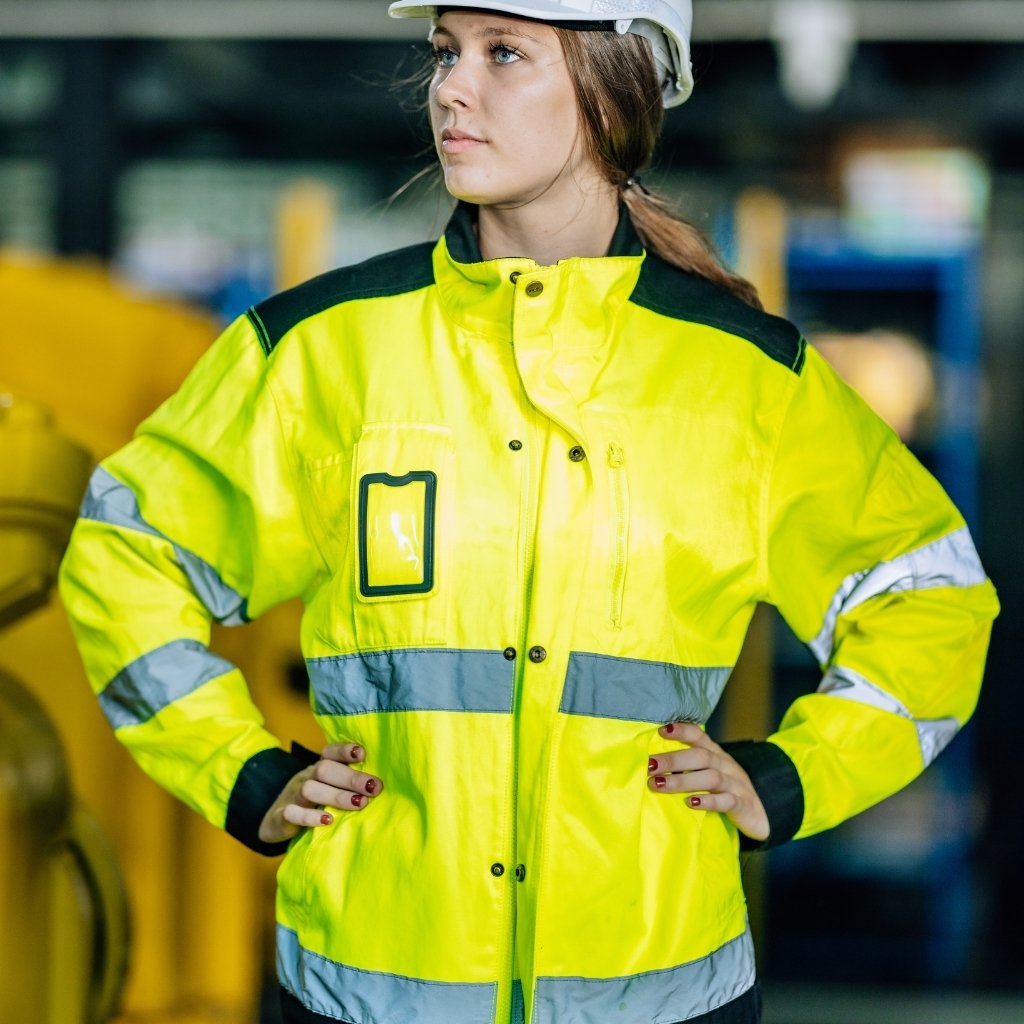 StormFlex Jacket: Waterproof, Durable, & Comfortable Rainwear 4 A young woman stands confidently in a bright yellow, high-visibility safety jacket with reflective stripes and black accents, wearing a white hard hat. Her hands rest on her hips in an industrial setting with large yellow machinery in the background.