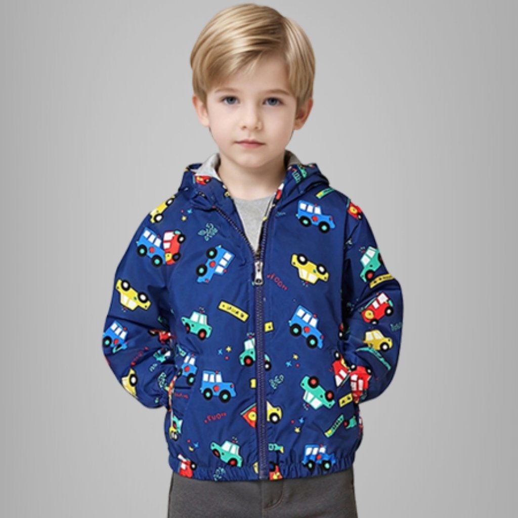 A young boy with blond hair and blue eyes stands facing forward, wearing a blue hooded jacket with a colorful pattern of cartoon cars. The jacket is zipped up, and his hands are in his pockets. He wears a gray T-shirt and gray pants, against a solid light gray background.