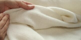 Double-Ply Grade A Cashmere Fabric Luxury Guide Close-up of a pale hand touching a soft, cream-colored knitted fabric. The fabric is fine-knit and draped in gentle folds, suggesting warmth and luxury; the background is blurred.