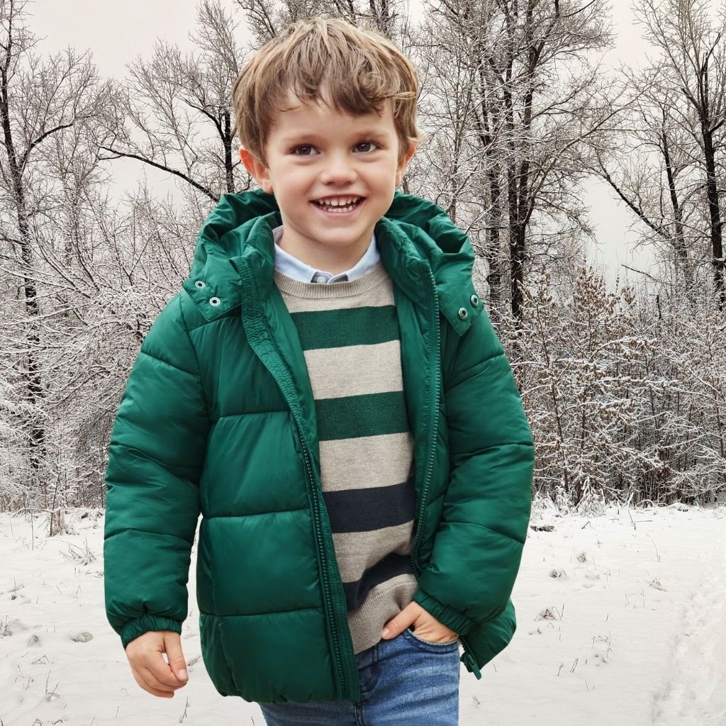 A young boy wearing a green down jacket stands in a snowy landscape with bare trees in the background.