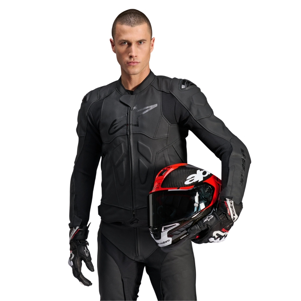 A man wearing a black Dusk leather motorcycle jacket and matching black pants holds a red and black helmet while standing against a plain white background.