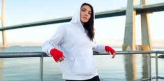 Fitted Hoodie: Sleek Style, Perfect Fit & Everyday Comfort A woman with dark hair wearing a gray hoodie and red hand wraps rests against a metal railing by a body of water, with a large bridge in the background.