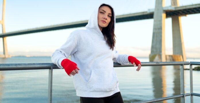 A woman with dark hair wearing a gray hoodie and red hand wraps rests against a metal railing by a body of water, with a large bridge in the background.