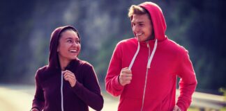 Fleece-Lined Zip Hoodie for Ultimate Winter Comfort Two people jogging side by side on a road. The person on the left wears a dark purple hoodie and smiles toward the person on the right, who wears a bright red hoodie. Both have hoods up and their arms bent as they run, with a blurred natural background of trees and road.