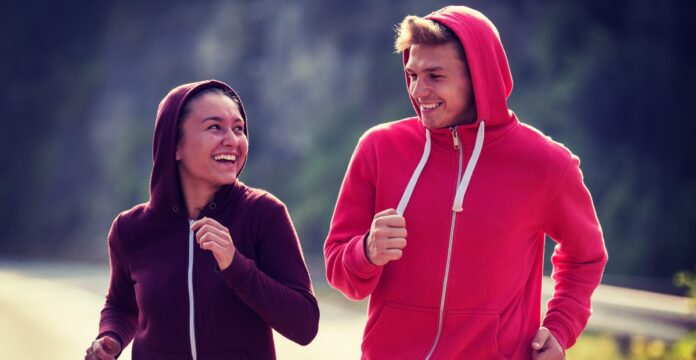 Two people jogging side by side on a road. The person on the left wears a dark purple hoodie and smiles toward the person on the right, who wears a bright red hoodie. Both have hoods up and their arms bent as they run, with a blurred natural background of trees and road.