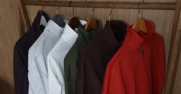 wooden clothing rack with multiple zip-up sweaters hanging on hangers, in colors including dark brown, gray, white, olive green, dark brown, and red, against a light brown paneled wall.