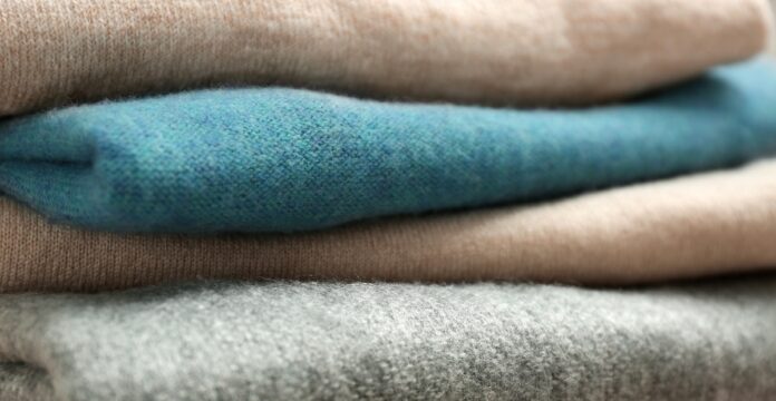 Heavyweight Pure Cashmere Fabric Close-up of a neat stack of folded heavyweight cashmere sweaters in beige, teal, beige, and gray, with prominent knit textures.