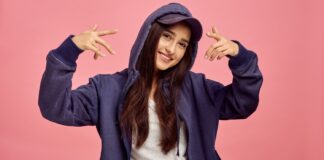 Hoodies with Anime: Trendy Styles for True Otakus A young woman with long dark hair smiles at the camera while wearing a dark blue zip-up hoodie with the hood up and a matching cap. She stands against a solid pink background, and her arms are raised with finger-gun gestures.