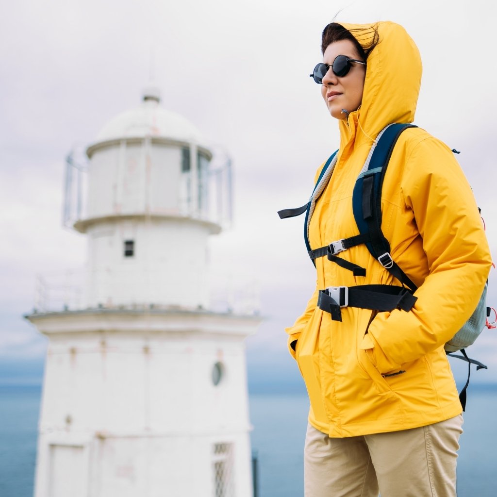 StormFlex Jacket: Waterproof, Durable, & Comfortable Rainwear 5 A woman wearing a bright yellow hooded jacket and round sunglasses stands with hands in pockets, looking toward the ocean. She has a backpack on, and a white lighthouse appears in the cloudy background.