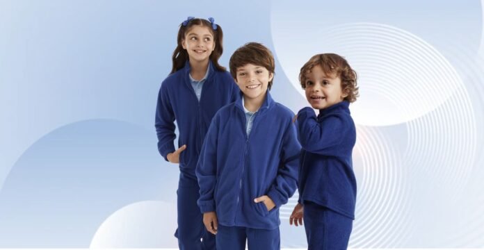 Three children wearing matching blue fleece jackets and pants, standing side by side against a blue and white abstract background; a girl with pigtails and blue bows on the left, a smiling boy in the middle, and a younger boy with curly hair on the right.