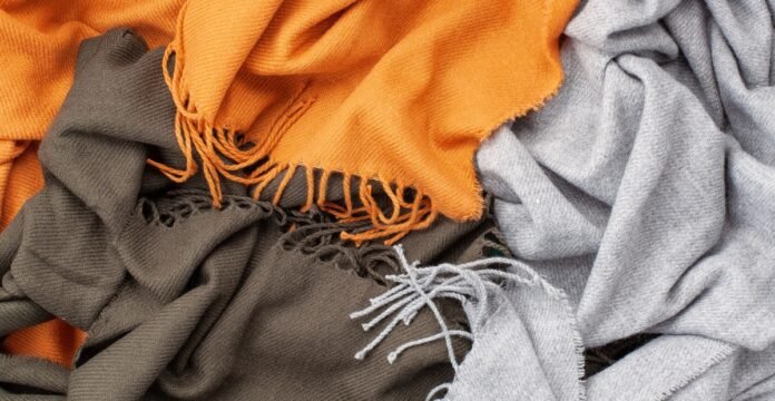 A close-up of a tangled pile of three scarves in orange, olive green, and light grey, showing woven textures and fringed edges.