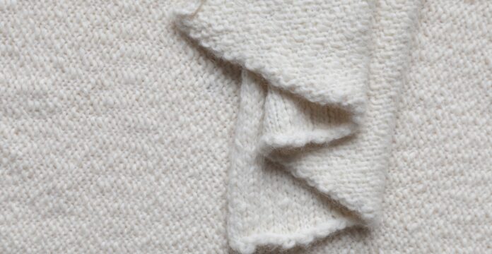 Lightweight Recycled Cashmere Fabric A close-up view of a cream-colored knitted fabric with a wavy, folded edge, showing the textured knit pattern.