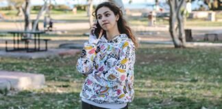 Logo Graphic Hoodie: Trendy Streetwear Style Guide A young woman stands in a park talking on her cellphone, wearing a white hoodie with colorful cartoon designs. Trees, grass, and picnic tables are in the background, with a hint of water in the distance.