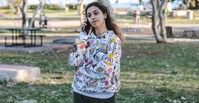 A young woman stands in a park talking on her cellphone, wearing a white hoodie with colorful cartoon designs. Trees, grass, and picnic tables are in the background, with a hint of water in the distance.