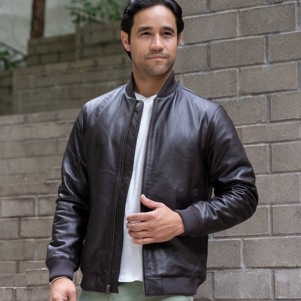Premium Shirt Style Leather Jacket for Casual and Formal Looks 2 A man stands against a light gray brick wall wearing a dark brown leather bomber jacket over a white T-shirt. He has dark hair and looks slightly to the side. The jacket is zipped up with one hand inside the pocket, and he wears light green pants and a silver bracelet.