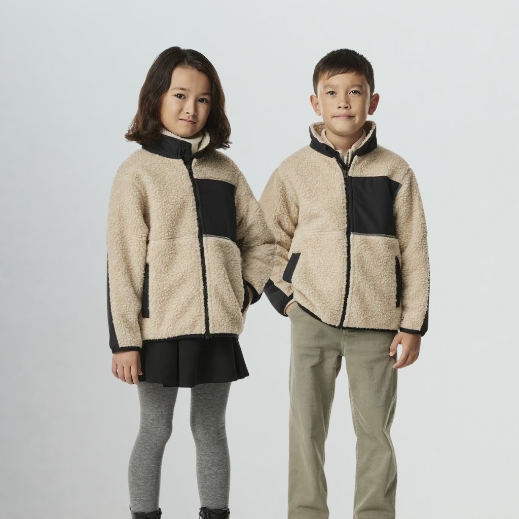 Two children, a girl and a boy, stand side by side in a studio wearing matching tan fleece jackets with black pockets and trim, against a plain white background. The girl wears a black skirt and gray tights with boots, while the boy wears olive pants; they stand close together with a relaxed pose.