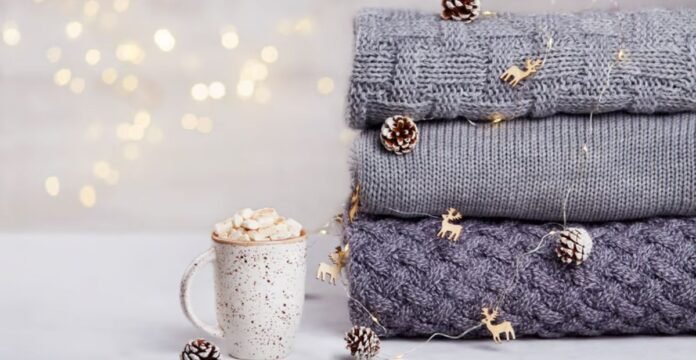 Alt text: A cozy winter scene showing a white speckled mug filled with marshmallows beside a stack of folded knit sweaters in gray and charcoal tones, with small decorative pinecones and string lights in the background.Winter Lock Fabric: