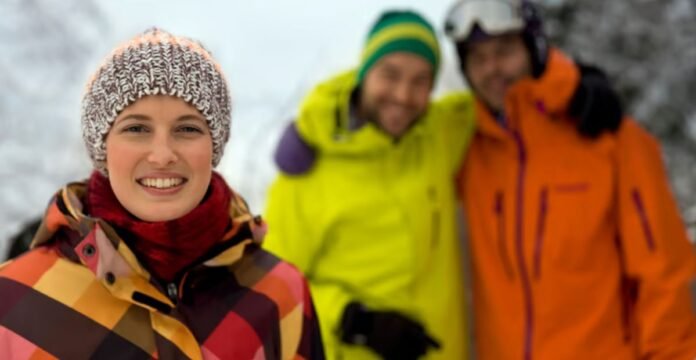Alt text: Group of three friends standing outdoors in a snowy landscape. The woman in the foreground wears a colorful plaid jacket and knit hat, smiling at the camera. Behind her, two men wearing bright yellow and orange winter jackets with hats and goggles smile as well; the background shows snowy trees and a wintry scene. Snow-Resistant Fabric Explained:
