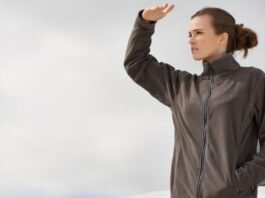 Alt text: A woman stands outdoors wearing a dark gray zip-up winter coat, shielding her eyes with her hand as she looks into the distance. What Is Wind Armor Extreme Fabric?