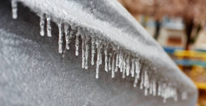 Alt text: Close-up of a gray, textured surface with many icicles hanging from its edge; a blurred background shows hints of a building and trees. Ice Barrier Windproof Fabric: