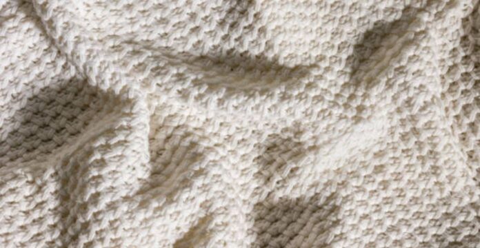 Alt text: Close-up of a cream-colored knit blanket with a dense, square pattern creating a three-dimensional texture; the blanket is draped and folded, casting soft shadows. Arctic Hug Comfort Weave: