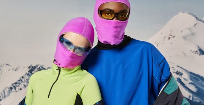 Alt text: Two models standing in front of snow-covered mountains. The left model wears a bright neon-green top and pink ski mask with light blue sunglasses; the right model wears a blue jacket over a black turtleneck, with a pink ski mask and yellow-tinted sunglasses.Polar Shield Windproof Knit Explained: