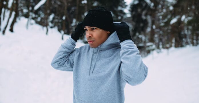 A man in a black beanie and gloves adjusts a gray hoodie in a snowy outdoor setting with snow-covered trees in the background.Snow Defense Performance Knit: