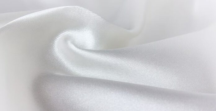 Close-up of soft, white satin fabric. The material is draped and folded, with gentle movement and a subtle sheen from diffused lighting. Snow Aura Premium Shield Fabric: