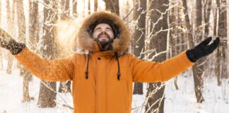 Heavyweight Insulated Winter Coat Explained: Features & Uses A man in a bright orange winter coat stands in a snowy forest with arms outstretched and a joyful expression, wearing a fur-lined hood. Heavyweight Insulated Winter Coat Explained: