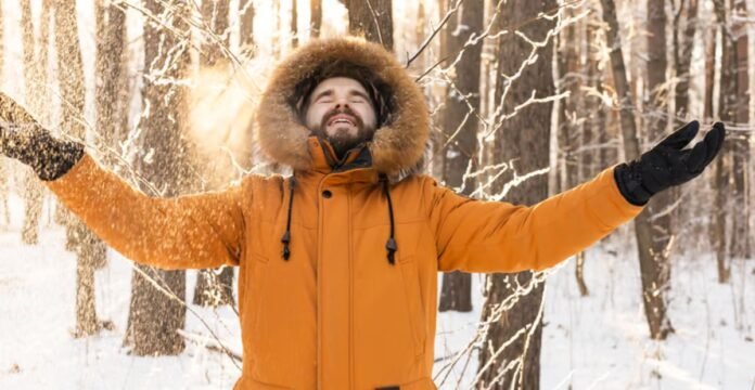 A man in a bright orange winter coat stands in a snowy forest with arms outstretched and a joyful expression, wearing a fur-lined hood. Heavyweight Insulated Winter Coat Explained: