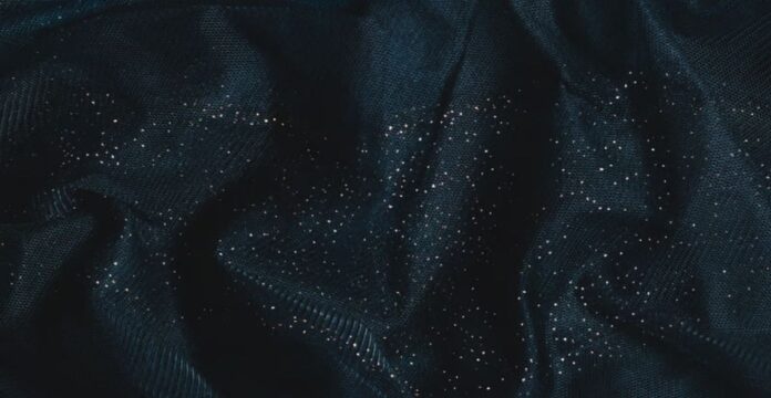 A close-up of dark blue sheer fabric with numerous small glittering silver speckles, gathered and folded to show texture and depth. Winter Glow Supreme Fabric: