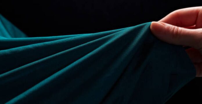 A close-up of teal fabric being held and draped by a hand, with soft folds and a dark background. Ultra Motion Stretch Fabric:
