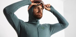 Power Dry Elite Knit Technology: Comfort Meets Performance A bearded man in a teal hooded long-sleeve zippered top pulls down ski goggles toward his eyes. He wears a glove-like sleeve with finger holes, and a small green starburst logo is on the chest. The background is a plain white wall. Power Dry Elite Knit Technology: