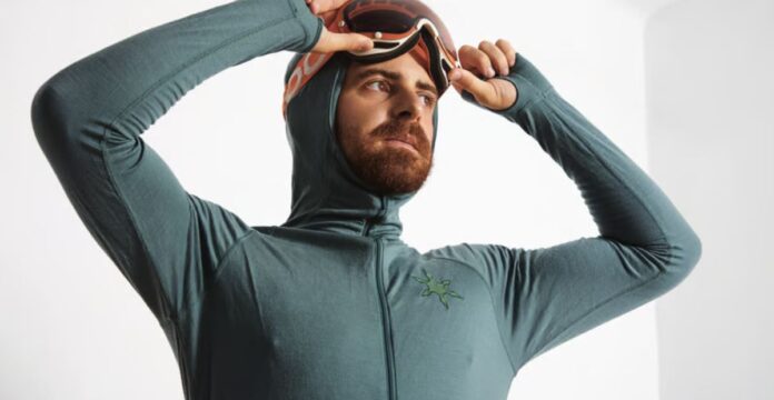A bearded man in a teal hooded long-sleeve zippered top pulls down ski goggles toward his eyes. He wears a glove-like sleeve with finger holes, and a small green starburst logo is on the chest. The background is a plain white wall. Power Dry Elite Knit Technology: