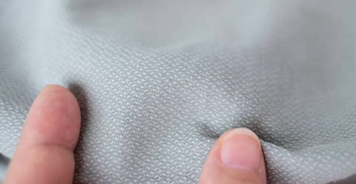 Close-up of light gray textured fabric being pinched by two fingers.Thermo Shield Performance Fabric: