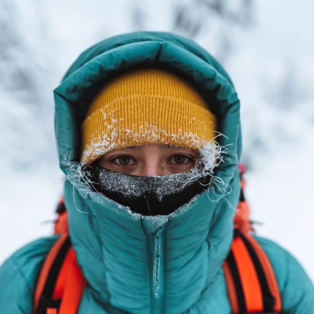 Alt text: Close-up of a person in winter gear with frost on the face: yellow beanie, teal puffer jacket with hood, and orange backpack straps. Only their eyes are visible through frost-covered fabric.

Snow-Resistant Fabric Explained: