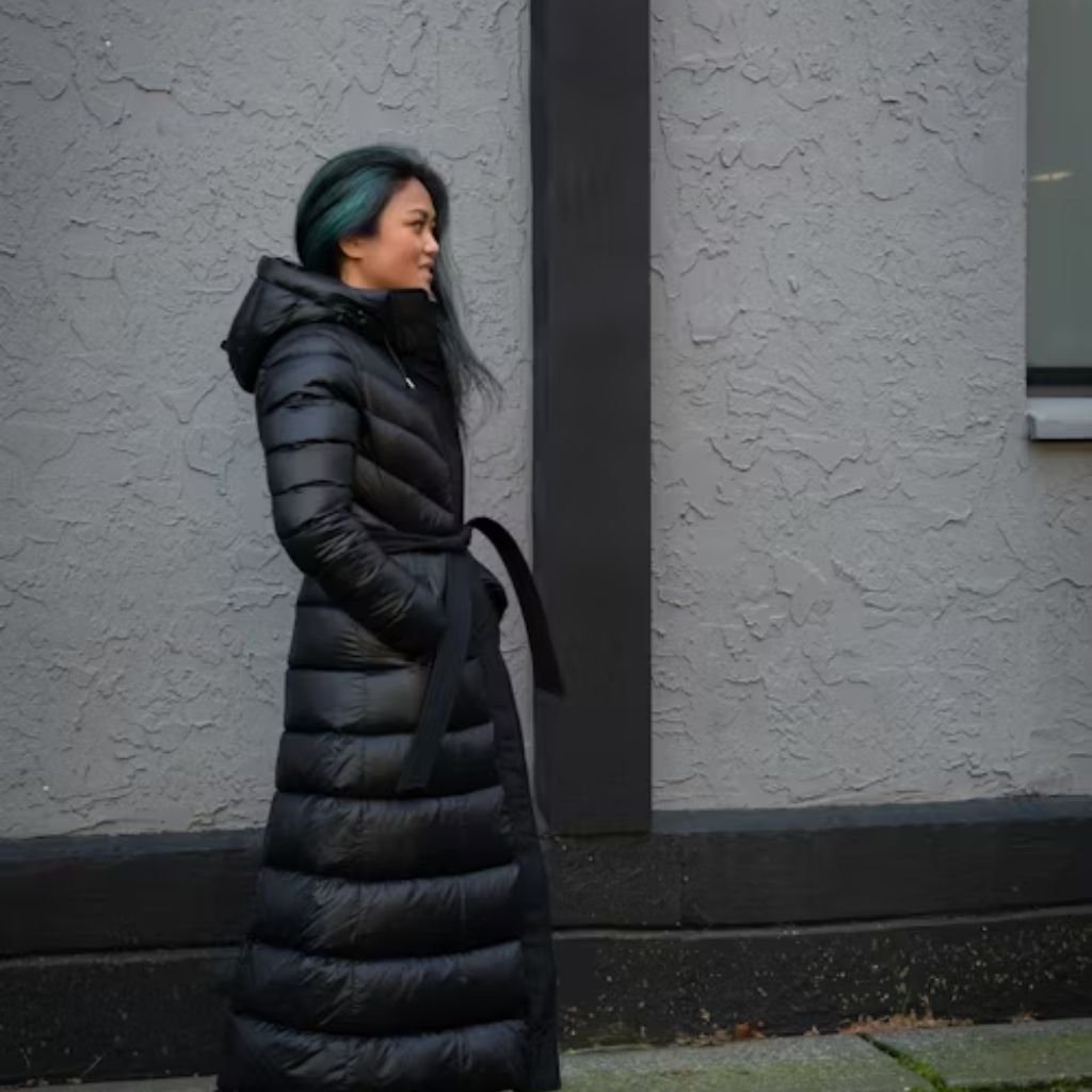 Cold Wind Blocking Winter Coat: Built to Stop Freezing Winds 8 Alt text (short description): A woman with teal and black hair wearing a long black puffer coat with a hood and belt, standing outdoors against a textured gray wall.
Cold Wind Blocking Winter Coat